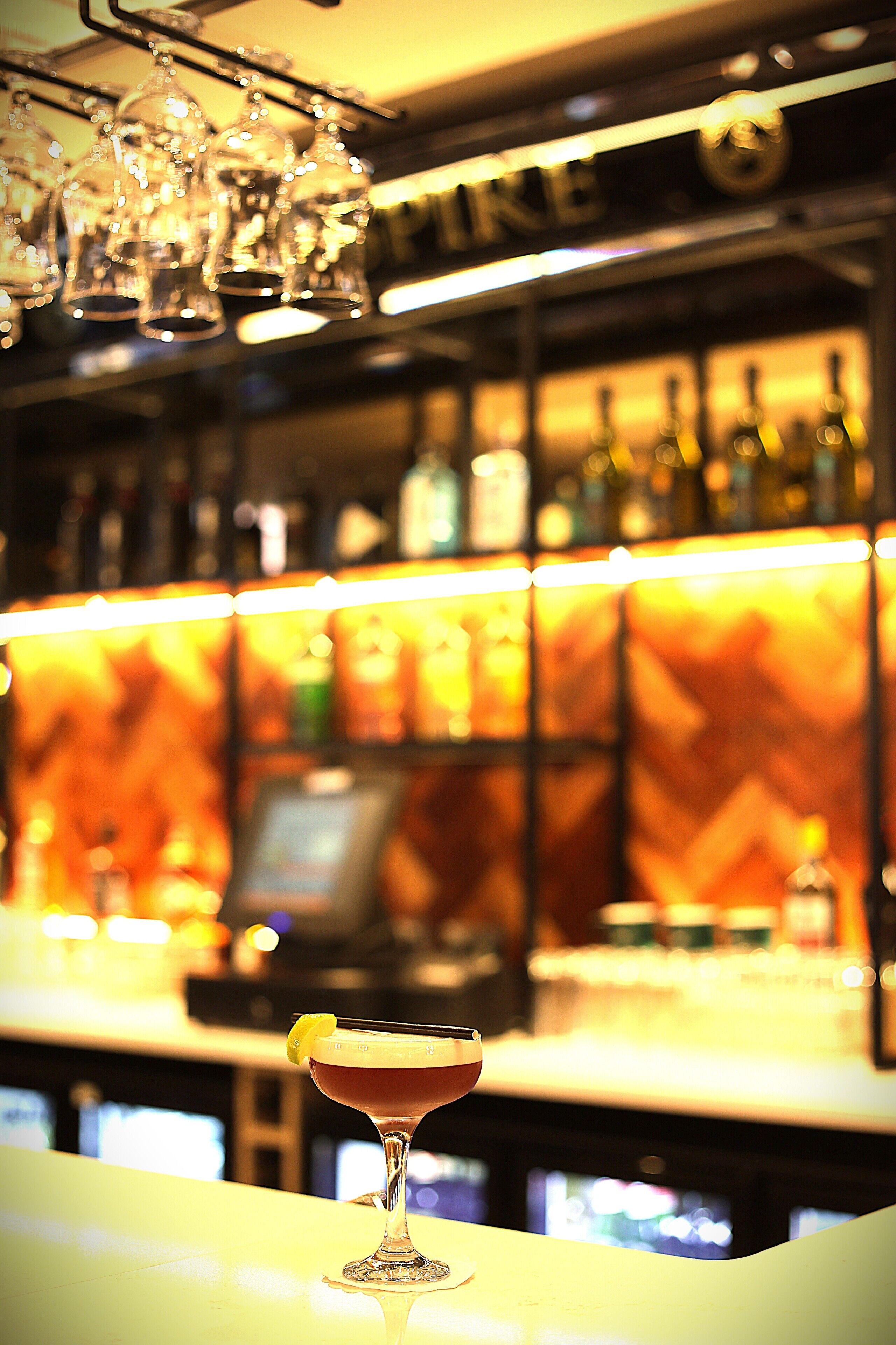 A bar with a glass of cocktail on the counter, shelves with bottles, and a warm, illuminated background.