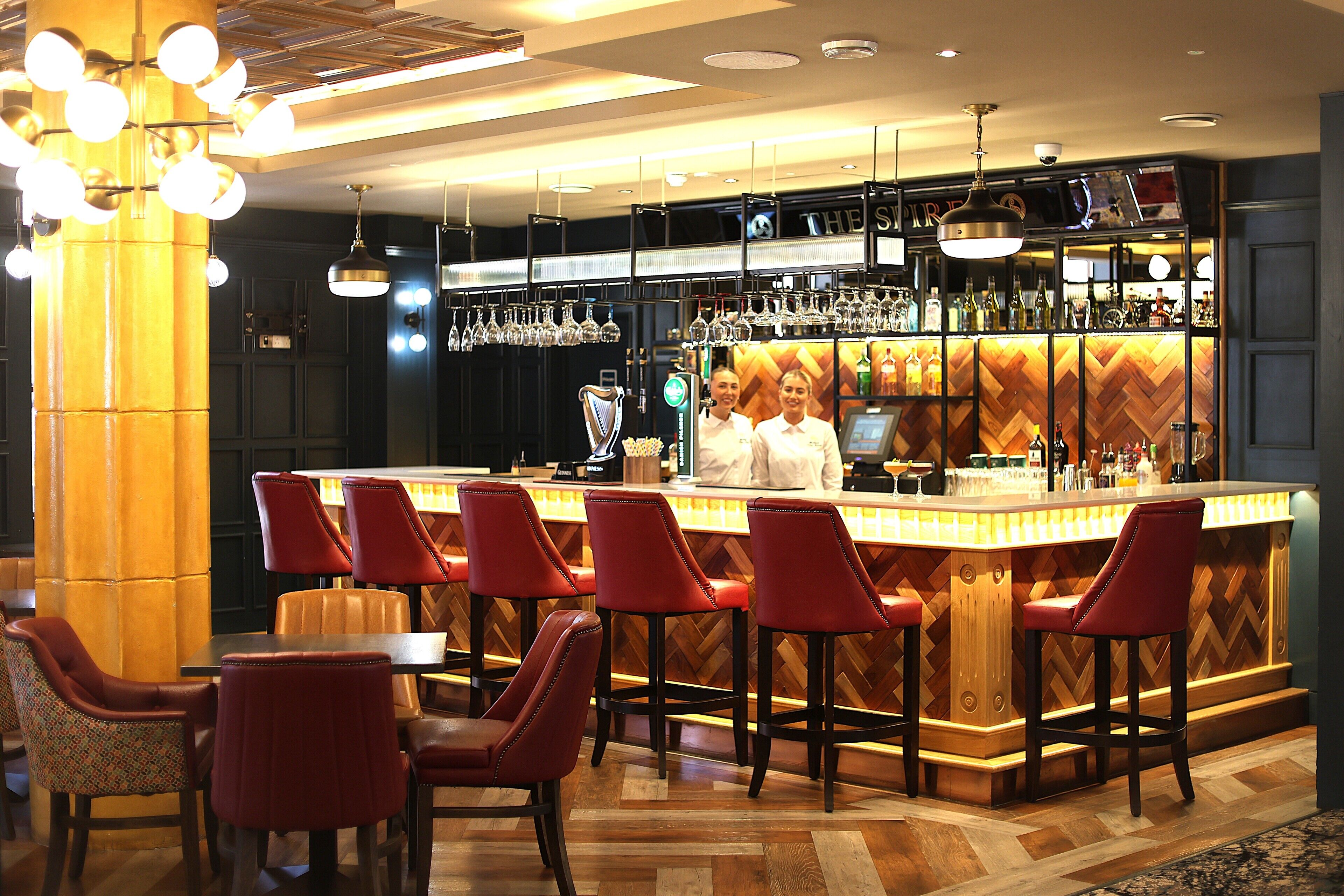 A bar area with red bar stools, a wooden herringbone floor, and a bar counter with a variety of bottles and glasses.