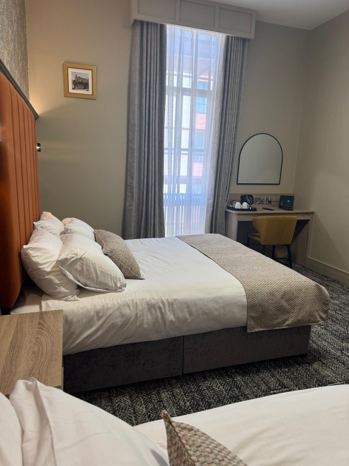 Ett hotellrum med en säng, ett skrivbord, en stol, en spegel och ett fönster med gardiner.