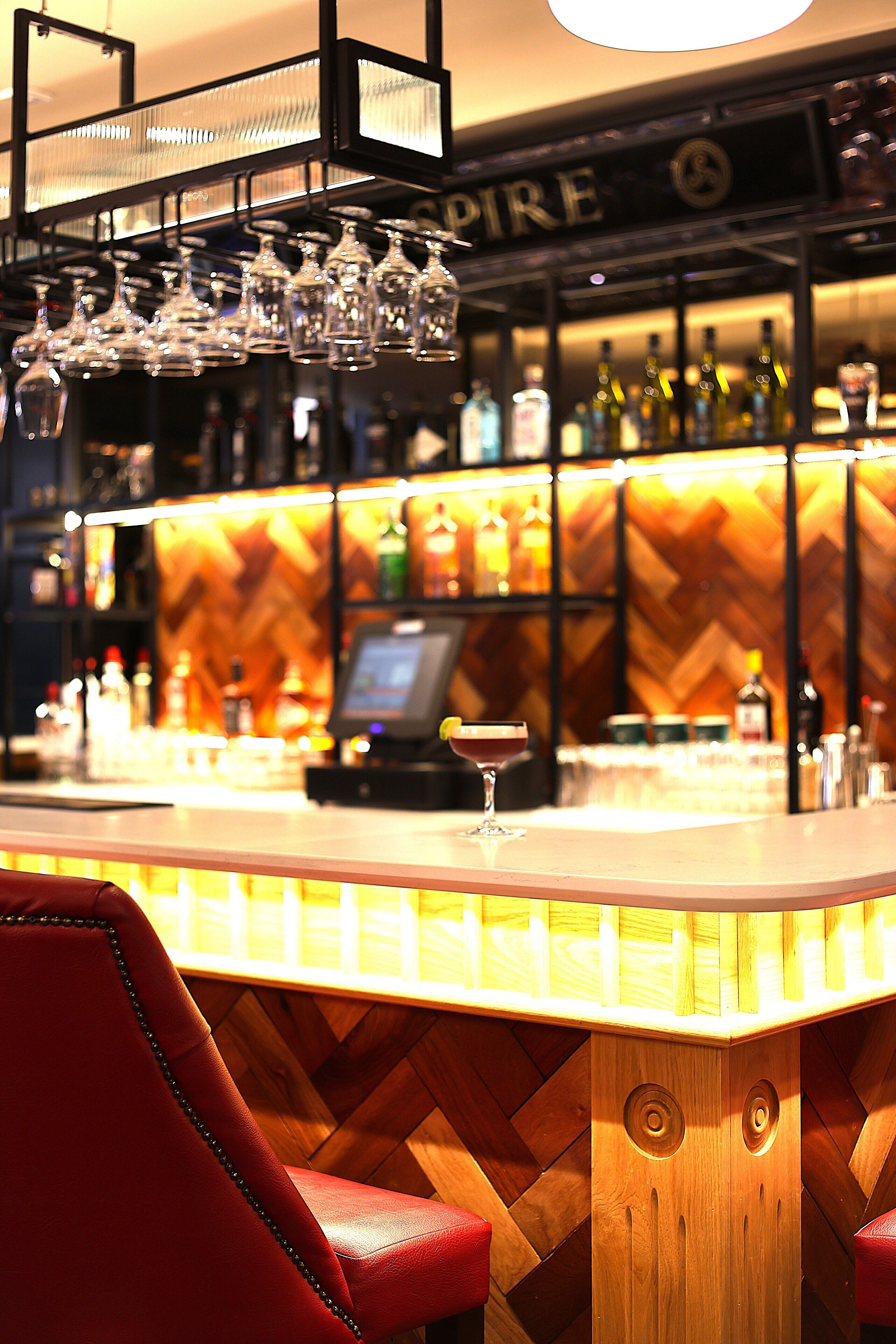 A bar area with a wooden herringbone wall, a red upholstered bar stool, and a variety of bottles and glasses on shelves.