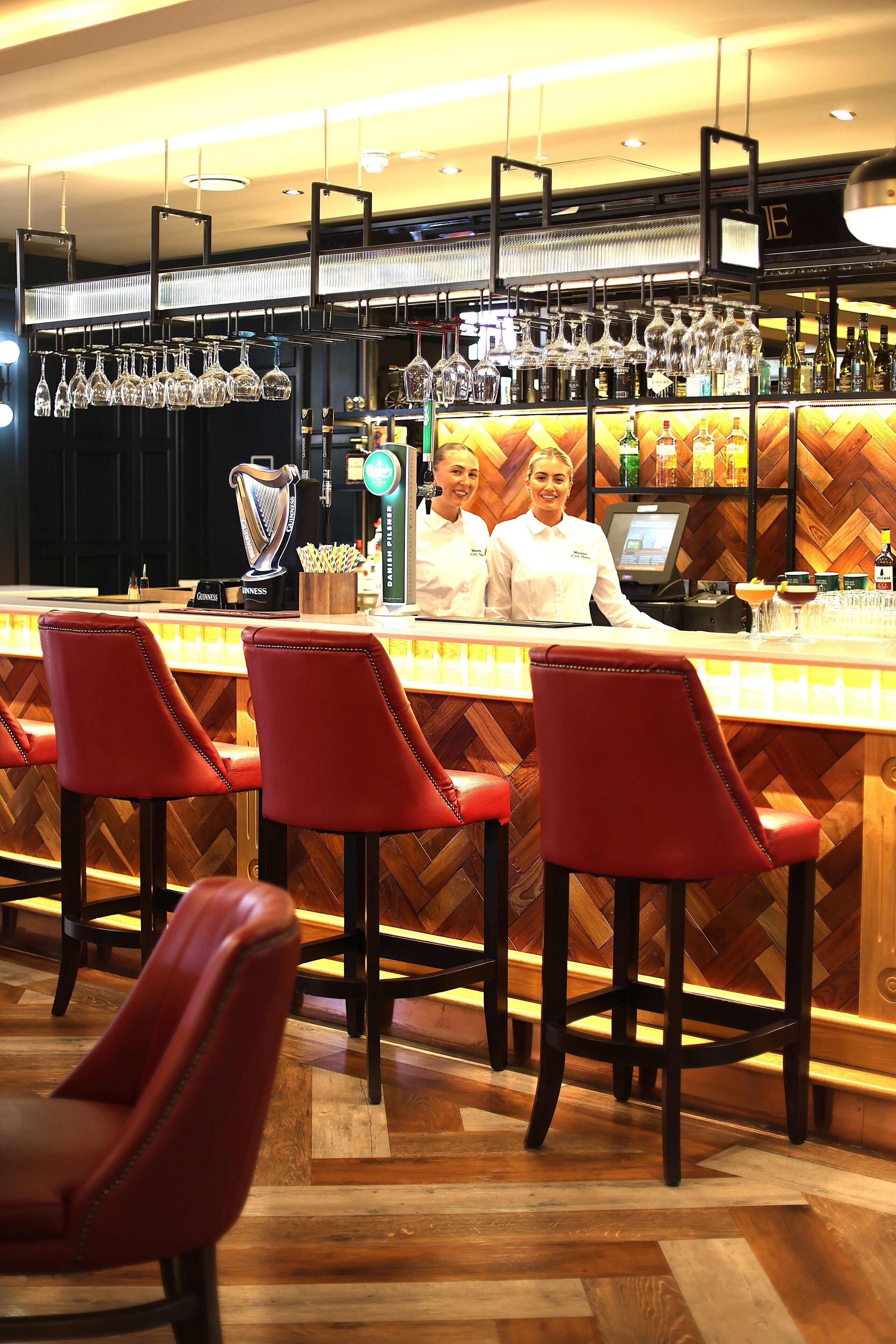 A bar area with red bar stools, a counter with a cash register, and a variety of bottles and glasses.