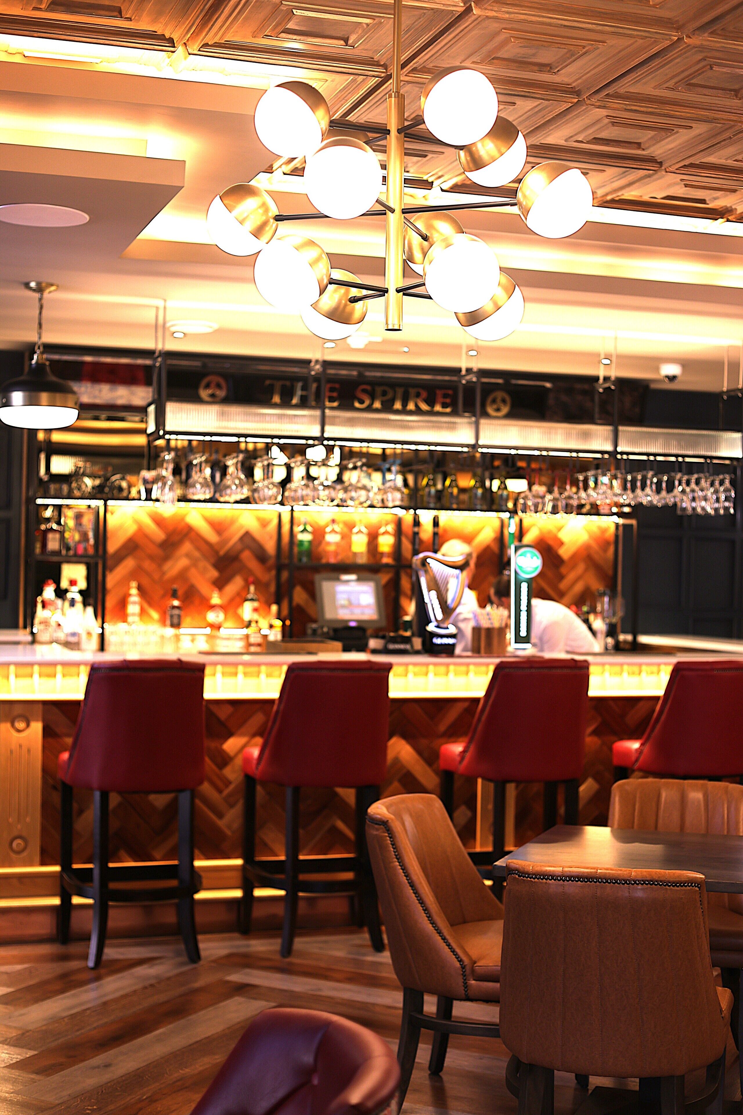 A bar area with red bar stools, a wooden herringbone floor, and a bar counter with various bottles and glasses.