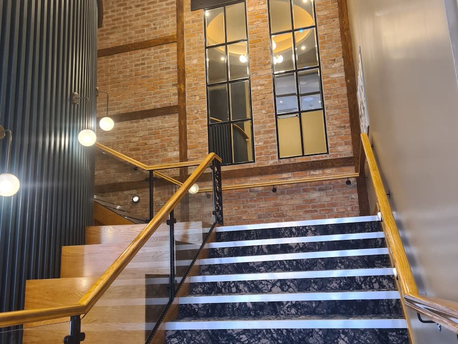 Una escalera con barandillas de madera y peldaños de mármol que conduce a una habitación con paredes de ladrillo y ventanas amplias.