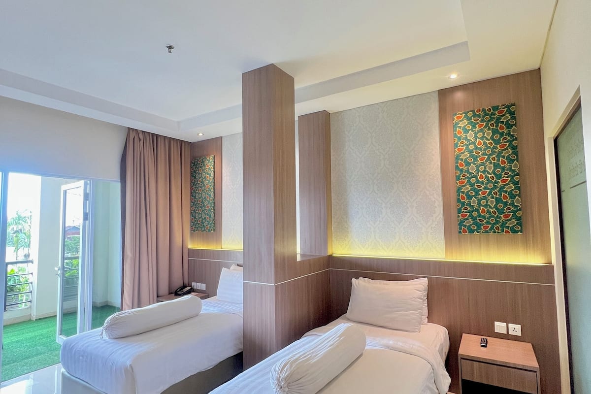 A hotel room with two beds, a small bedside table, a large window with curtains, and a wall with a decorative pattern.