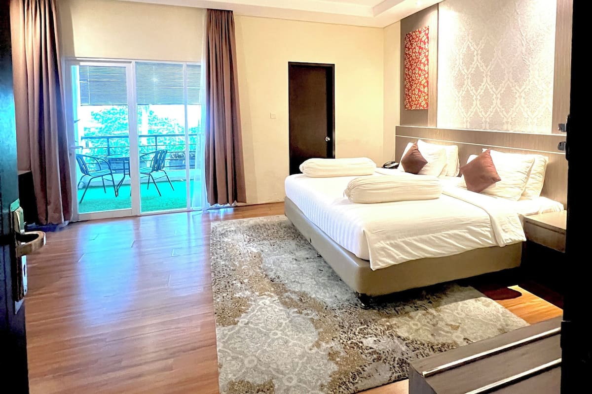 A modern hotel room with a large bed, a wooden floor, and a view of the outdoors through a sliding glass door.