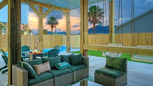 An outdoor patio with wicker furniture, a swing, and a view of a mountain and palm trees at sunset. - Pool, Firepit + Golf Cart Zone + Near Beach | Looney Dunes (Port Aransas)