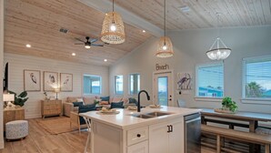 A spacious kitchen with a central island, wooden ceiling, pendant lights, and a view of the beach through the windows. - Pool, Firepit + Golf Cart Zone + Near Beach | Looney Dunes (Port Aransas)