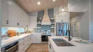 A modern kitchen with white cabinetry, a central island with a sink, stainless steel appliances, and a wooden ceiling. - Pool, Firepit + Golf Cart Zone + Near Beach | Looney Dunes (Port Aransas)