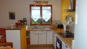 Une cuisine compacte équipée d’un évier, d’une cuisinière et d’un four. On y trouve une fenêtre avec des rideaux, ainsi qu’une petite table sur laquelle sont posés un panier et une machine à café.