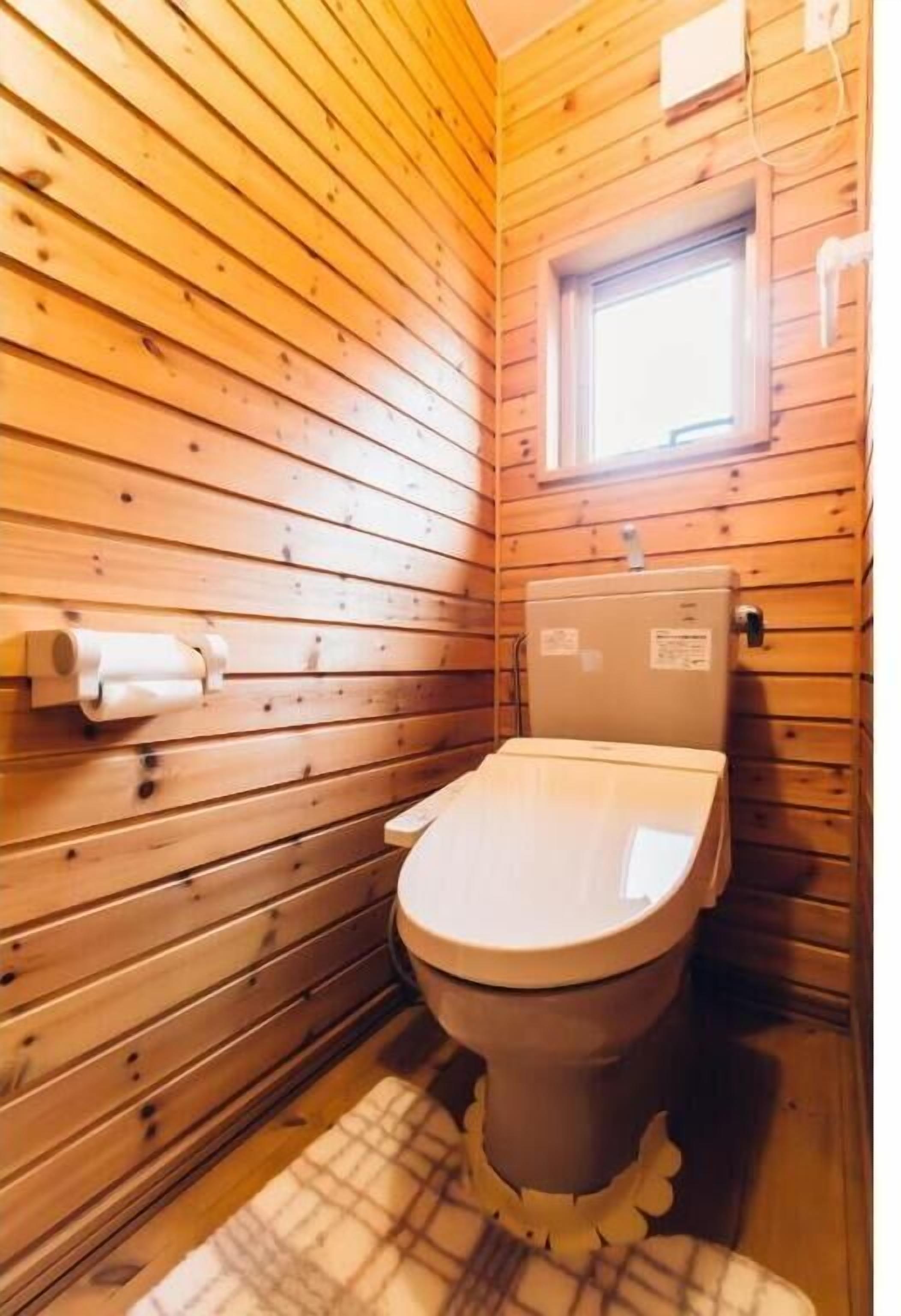 A small, wooden-paneled bathroom with a toilet, a window, and a towel dispenser.