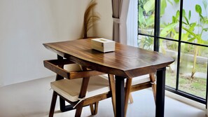 A wooden dining table with a box on top, a cushioned chair, and a view of greenery through a window. - Malasama suites hotel (Kuta)