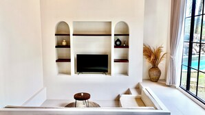 A living room with a flat-screen TV mounted on a white wall, surrounded by shelves with decorative items. - Malasama suites hotel (Kuta)