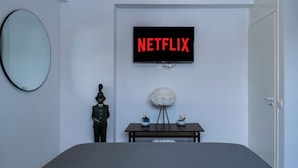 Un dormitorio con una cama, un televisor de pared mostrando "NETFLIX", un espejo redondo, una pequeña estatua y una silla blanca redondeada.