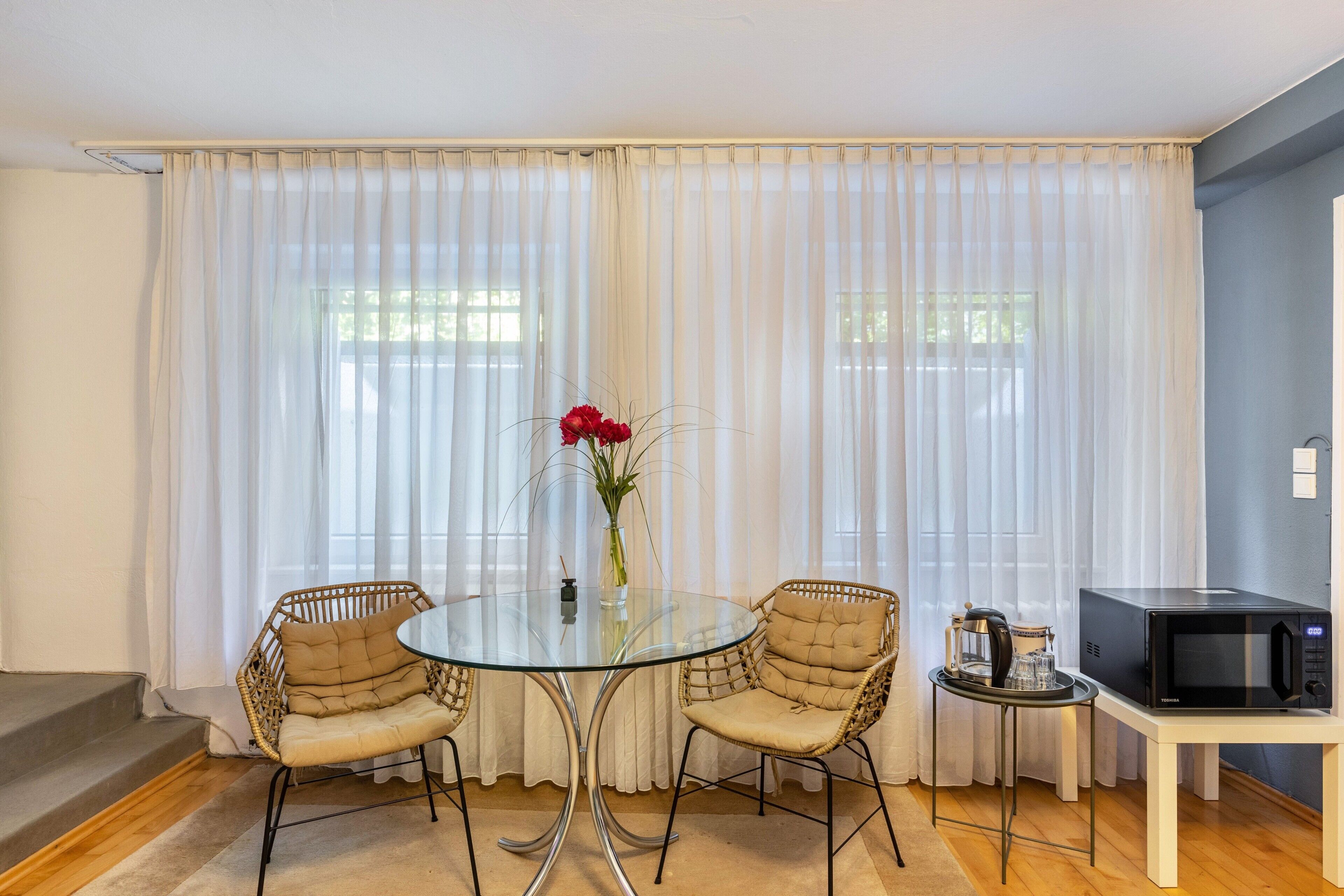 Ein Zimmer mit einem runden Glastisch, zwei Korbstühlen, einer Vase mit roten Blumen, einem kleinen Tisch mit einer Kaffeemaschine und einer Mikrowelle auf einem weißen Tisch.