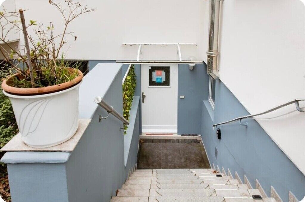 Eine enge Treppe führt zu einer Tür, einem Blumentopf und einem kleinen Fenster mit einem Schild.