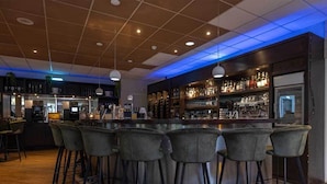 Zona de bar con barra circular y seis taburetes, mostrador con caja registradora y estanterías con botellas y vasos.