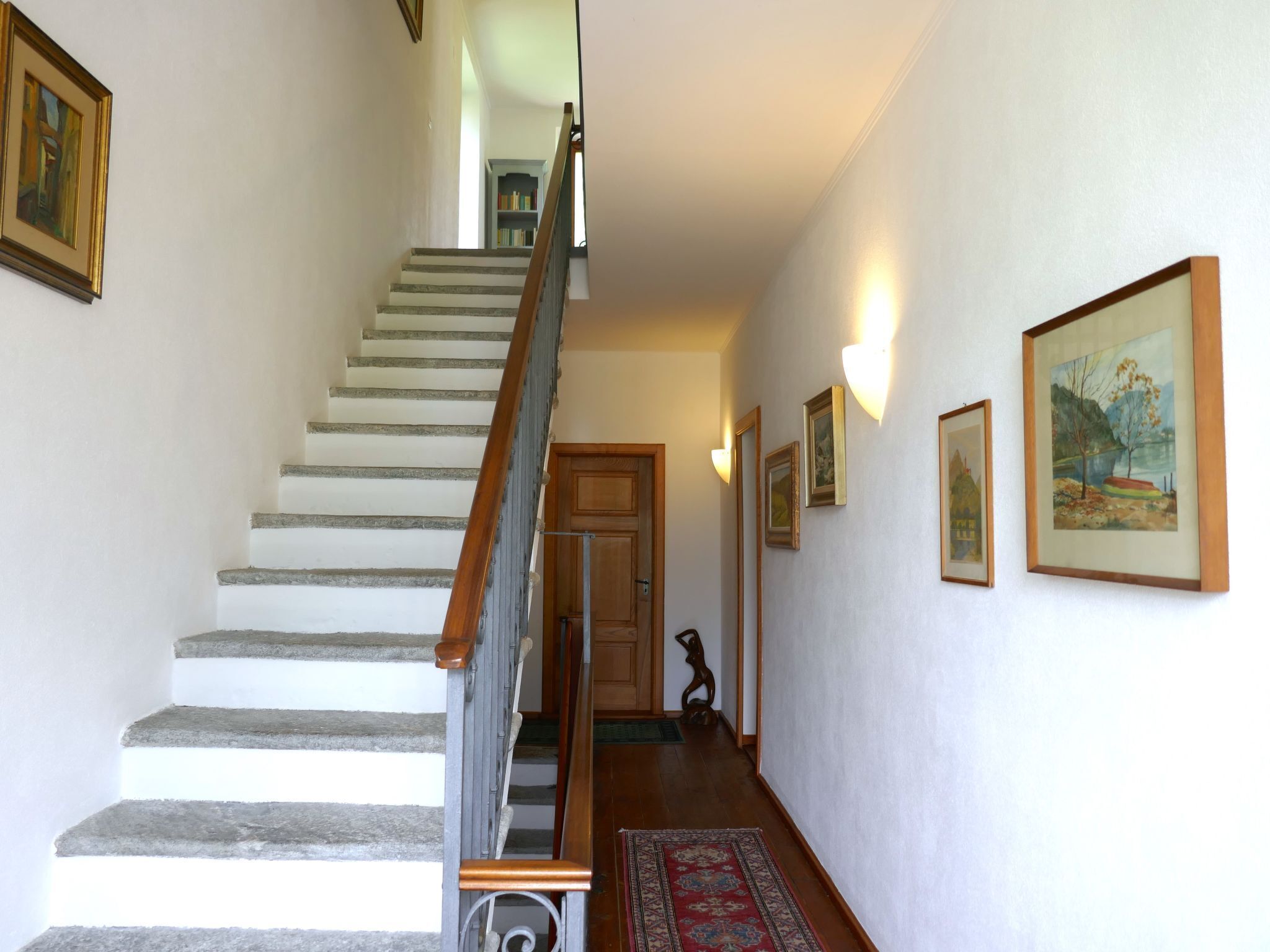 Un escalier avec des rampes en bois et des murs blancs, menant à une pièce dotée d’une porte et d’un tapis sur le sol.
