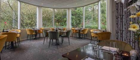 A restaurant with round tables and chairs, large windows with a view of trees, and a carpeted floor.