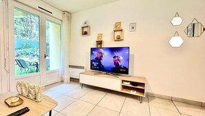 A living room with a TV, wooden furniture, a dining table, and a view of the outdoors. - Le Cosy Clim Jardin Parking Privé Tram 20min Plage (Montpellier)