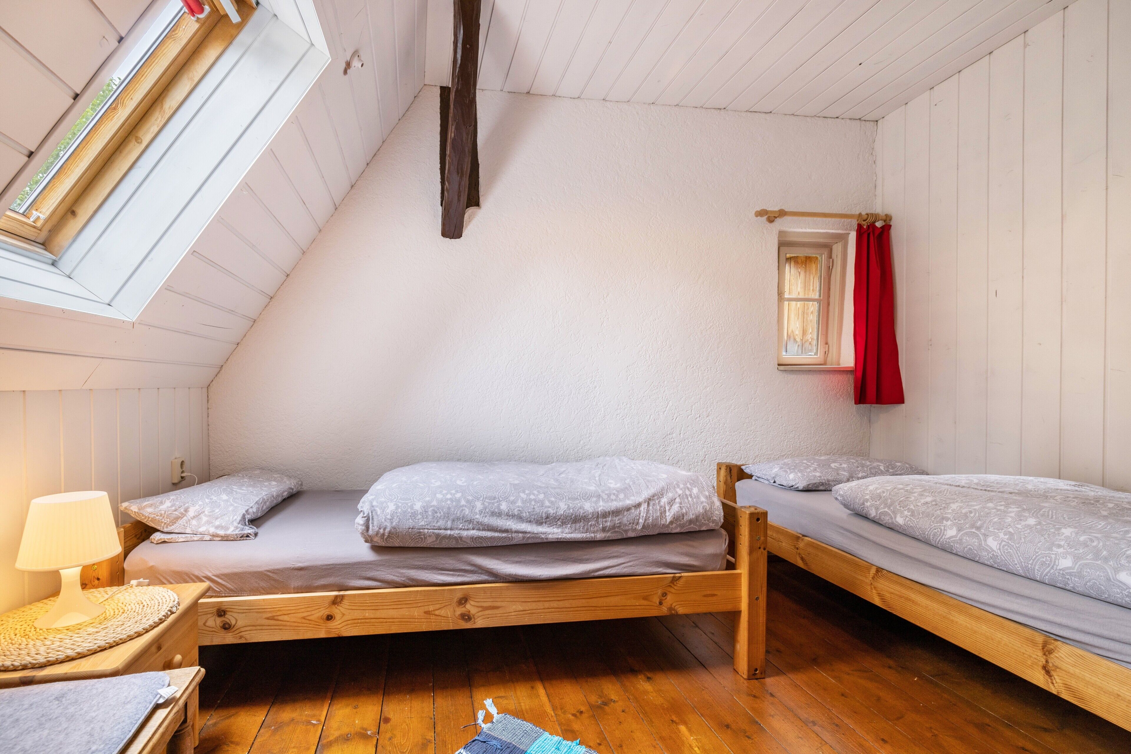 Ein kleiner, gemütlicher Raum mit zwei Holzbettgestellen übereinander, einem Fenster mit rotem Vorhang und einer Lampe auf einem Nachttisch.