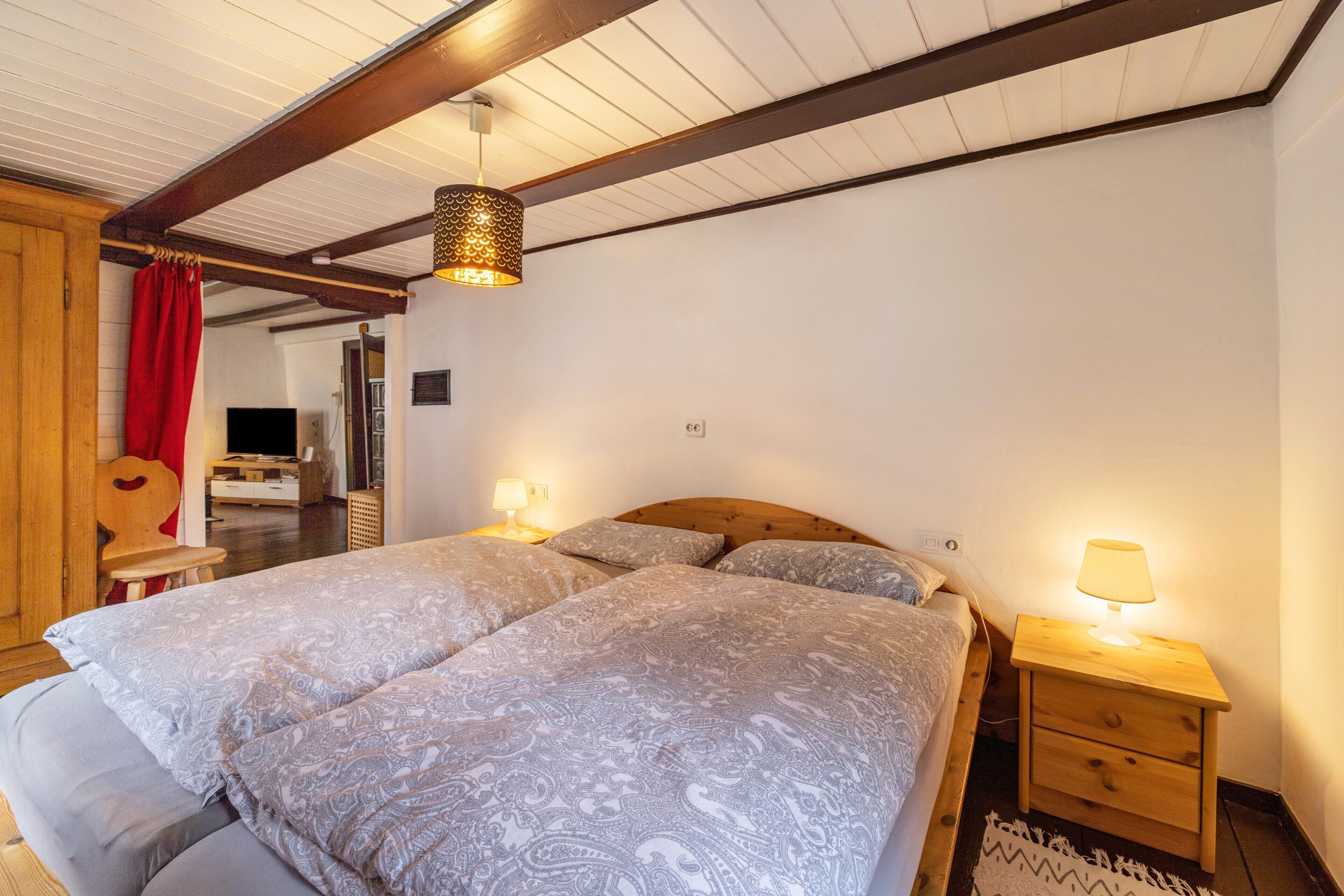Une chambre avec un plafond en bois, deux lits avec une literie à motifs, une table de chevet avec une lampe et un rideau rouge.
