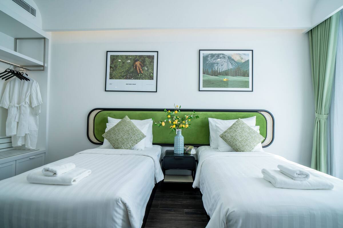 Ein Hotelzimmer mit zwei Betten, einem grünen Kopfteil, weißen Bettwäsche, einer Vase mit Blumen und zwei gerahmten Bildern an der Wand.