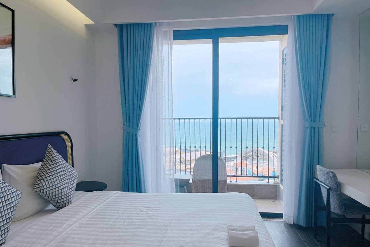 Ein Hotelzimmer mit Bett, Stuhl, einem kleinen Tisch und einem Balkon mit Meerblick.