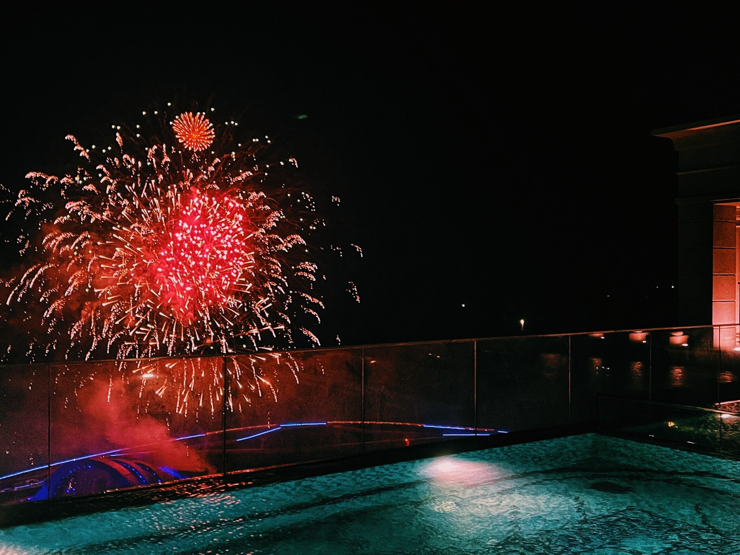 Vista nocturna de fuegos artificiales sobre el agua, con un edificio al fondo.