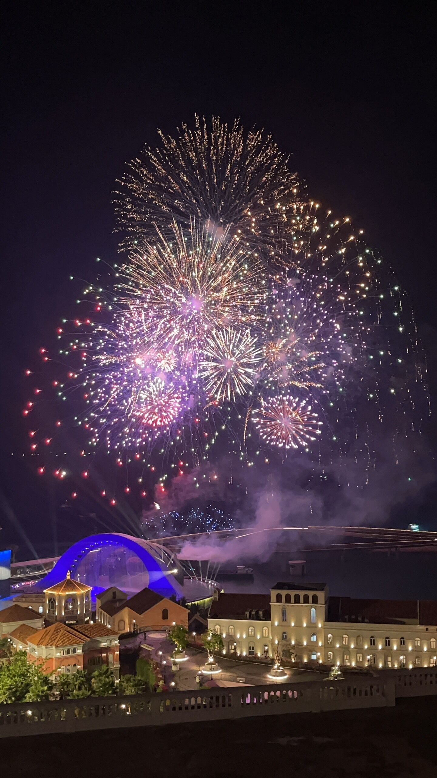 Vista noturna de uma orla com espetáculo de fogos de artifício, edifícios com fachadas iluminadas e uma estrutura em forma de cúpula.
