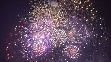 Vista noturna de uma orla com espetáculo de fogos de artifício, edifícios com fachadas iluminadas e uma estrutura em forma de cúpula.