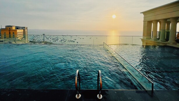 Una piscina infinita con vistas a la puesta de sol sobre el océano, un edificio con columnas y un horizonte urbano distante.