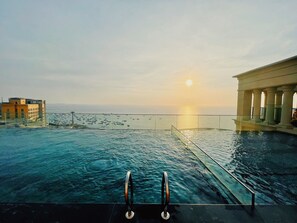 Una piscina infinita con vistas a la puesta de sol sobre el océano, un edificio con columnas y un horizonte urbano distante.