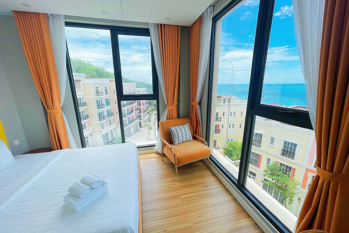 Ein Hotelzimmer mit großem Fenster, einem Bett, einem Sessel und Blick auf Gebäude und das Meer.