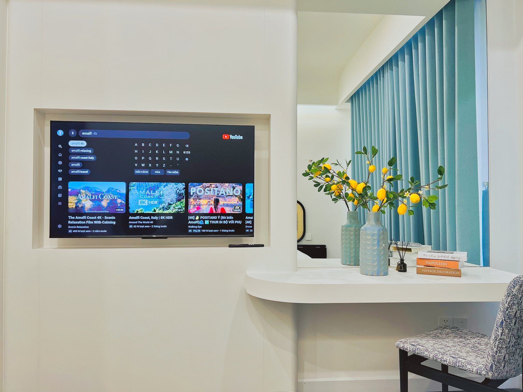 Una sala de estar moderna con un televisor montado mostrando un canal de YouTube, un estante con objetos decorativos y una ventana con cortinas azules.