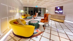 Un vestíbulo con piso a cuadros, una pantalla grande mostrando 'The Hill', y arreglos de asientos modernos que incluyen un sillón amarillo y una butaca naranja.