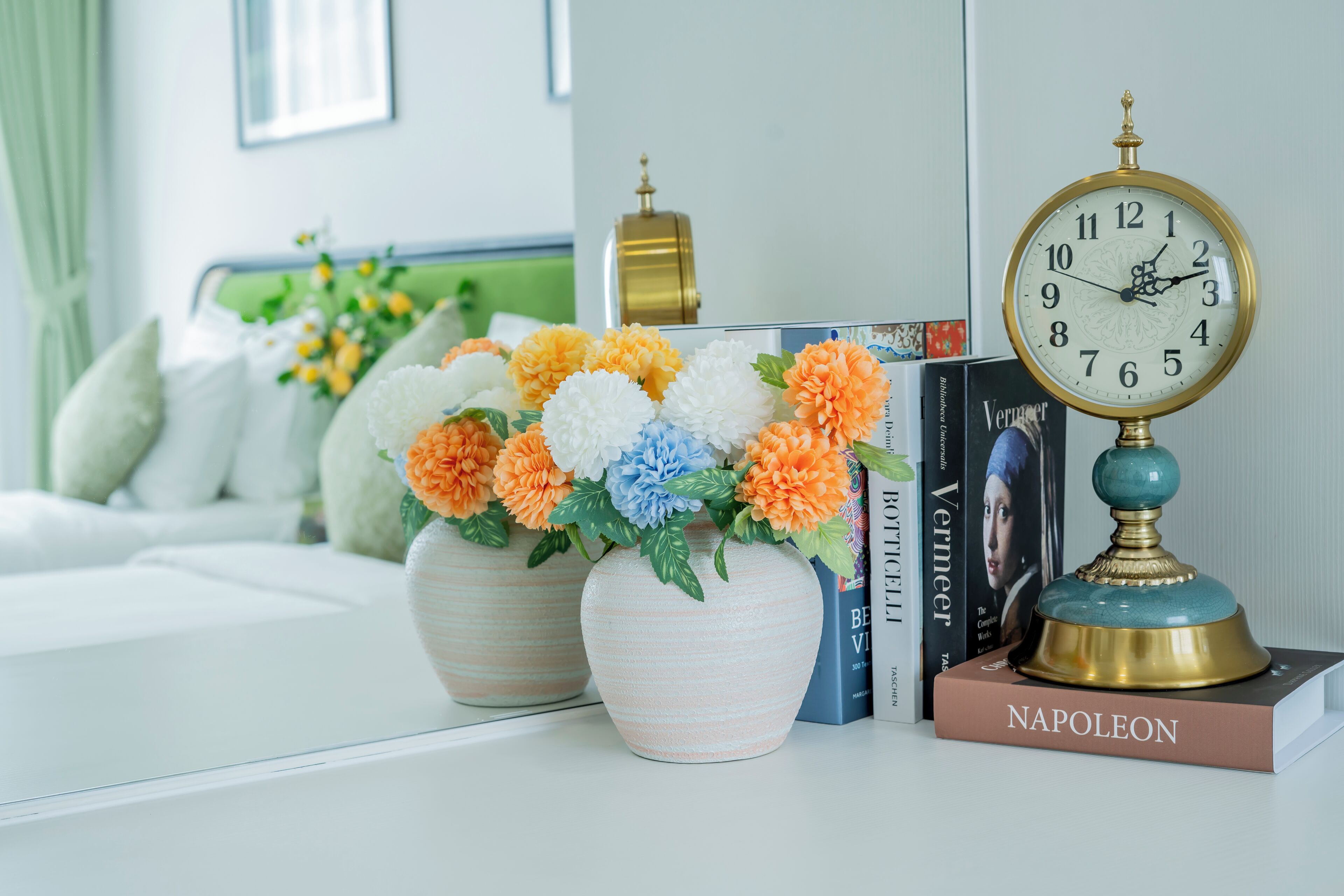 A white table with two vases of flowers, a clock, and books.