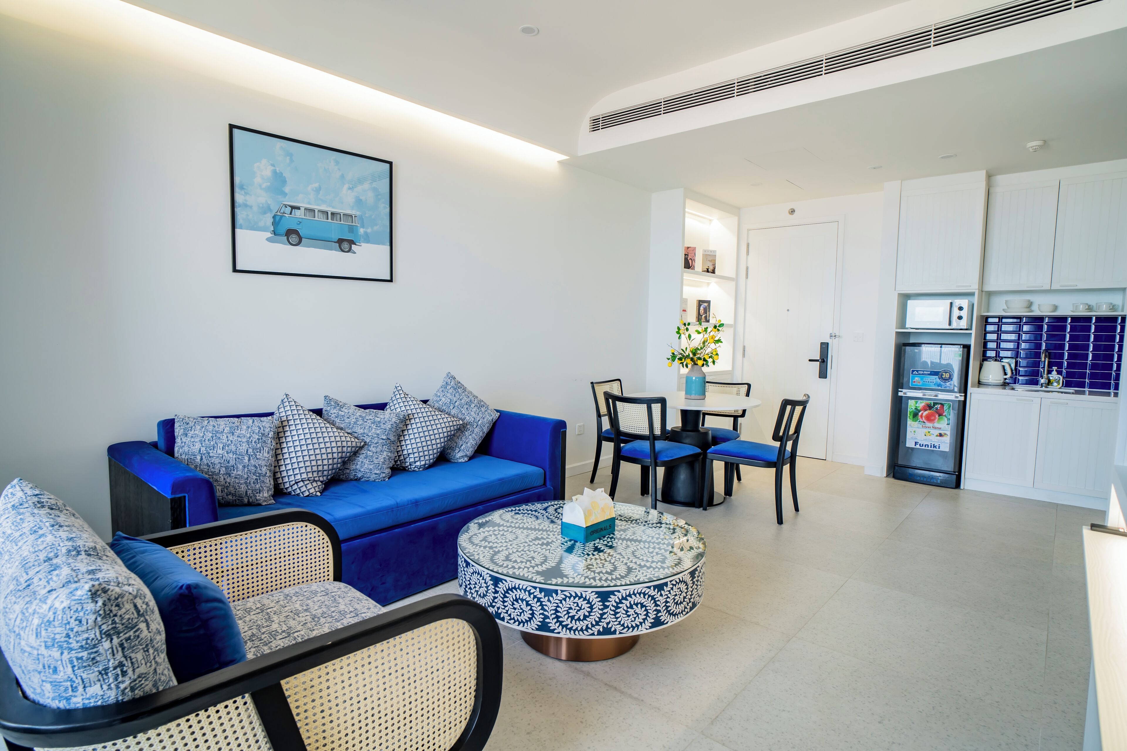 A modern living room with a blue sofa, round coffee table, and wicker chairs. A dining area with a table and chairs is visible in the background.