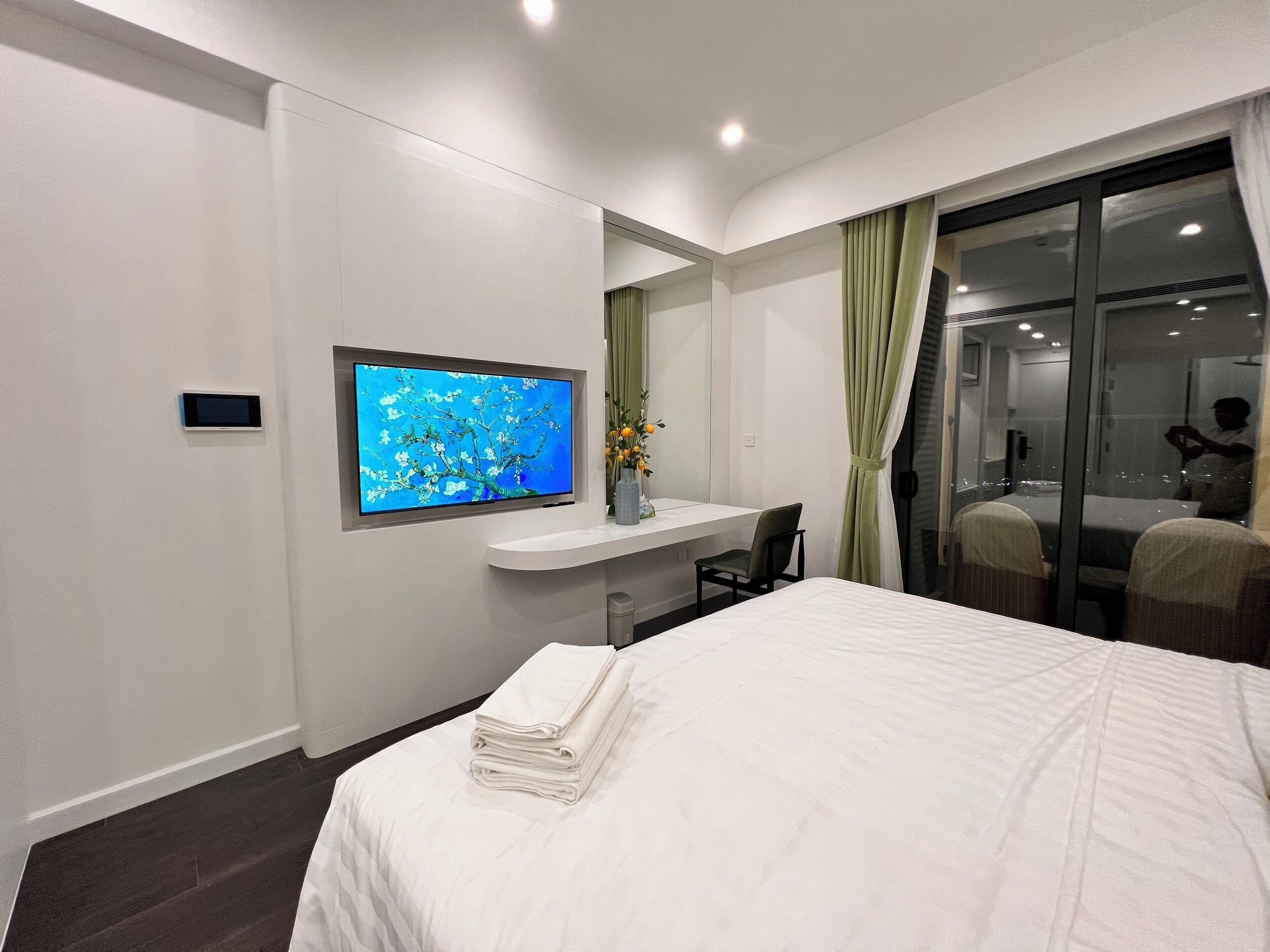 A modern hotel room with a bed, a TV mounted on the wall, a small desk, and a window with curtains.