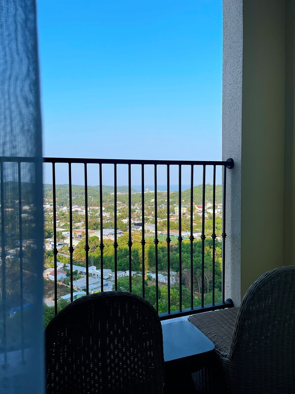 Un balcón con barandilla de metal, muebles de mimbre y vistas a un pueblo con zonas verdes y edificios.