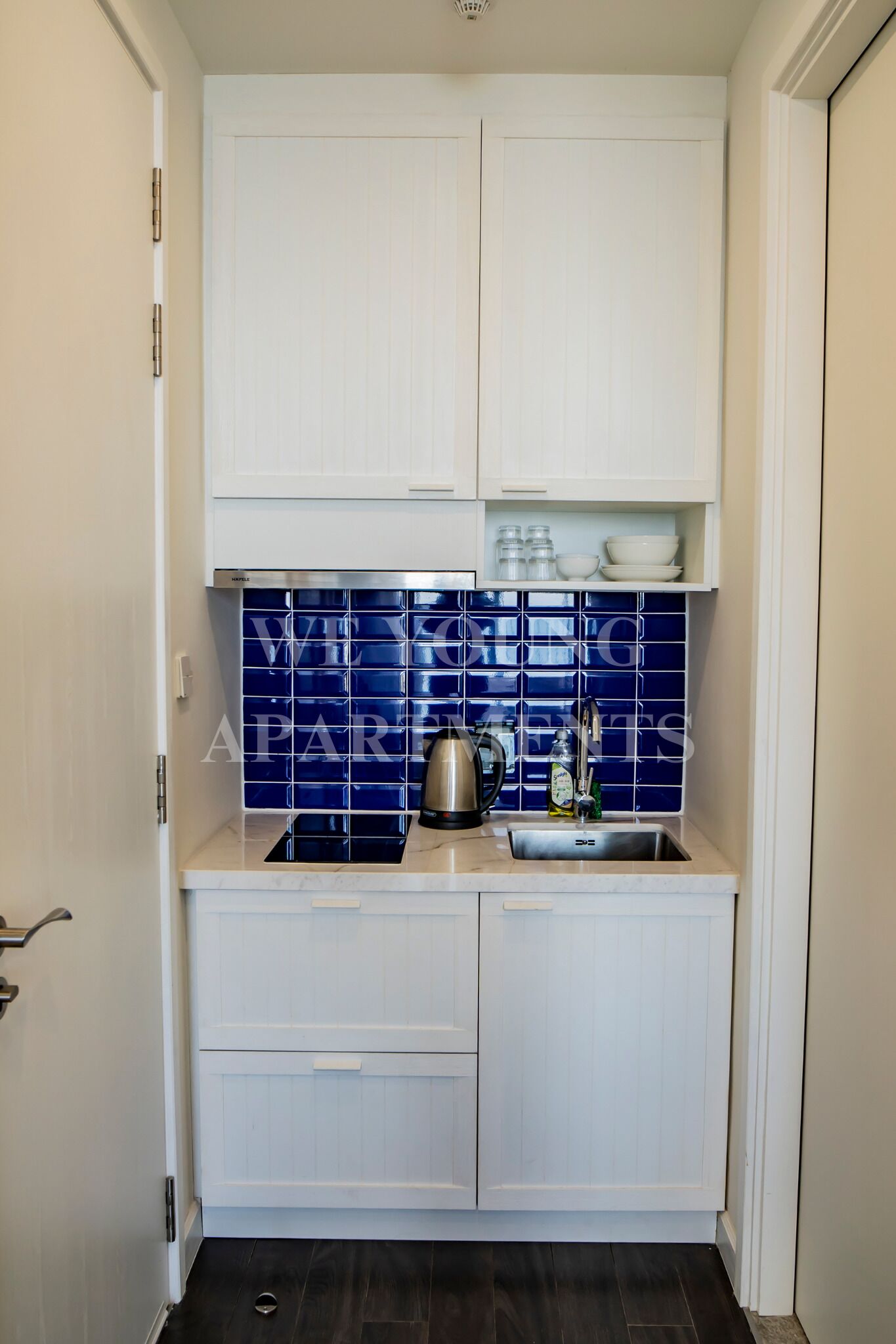 A compact kitchen with white cabinetry, a blue tiled backsplash, and a stainless steel kettle on the countertop.