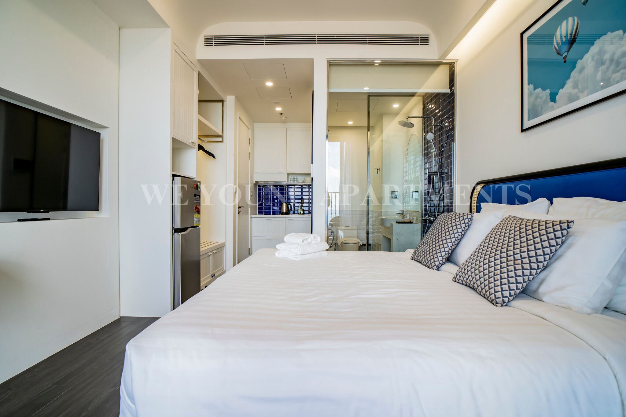 A modern hotel room with a large bed, a flat-screen TV, a kitchenette, and a glass shower area.