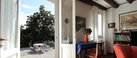 En åben dør ind til en terrasse med et bord og stole, en reol fyldt med bøger, og et maleri på væggen.