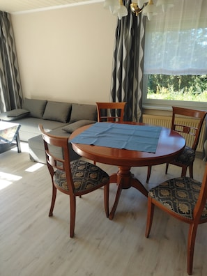 Ein Essbereich mit einem runden Holztisch, vier Stühlen mit gemusterten Sitzbezügen, einem grauen Sofa und Blick ins Grüne durch ein Fenster.