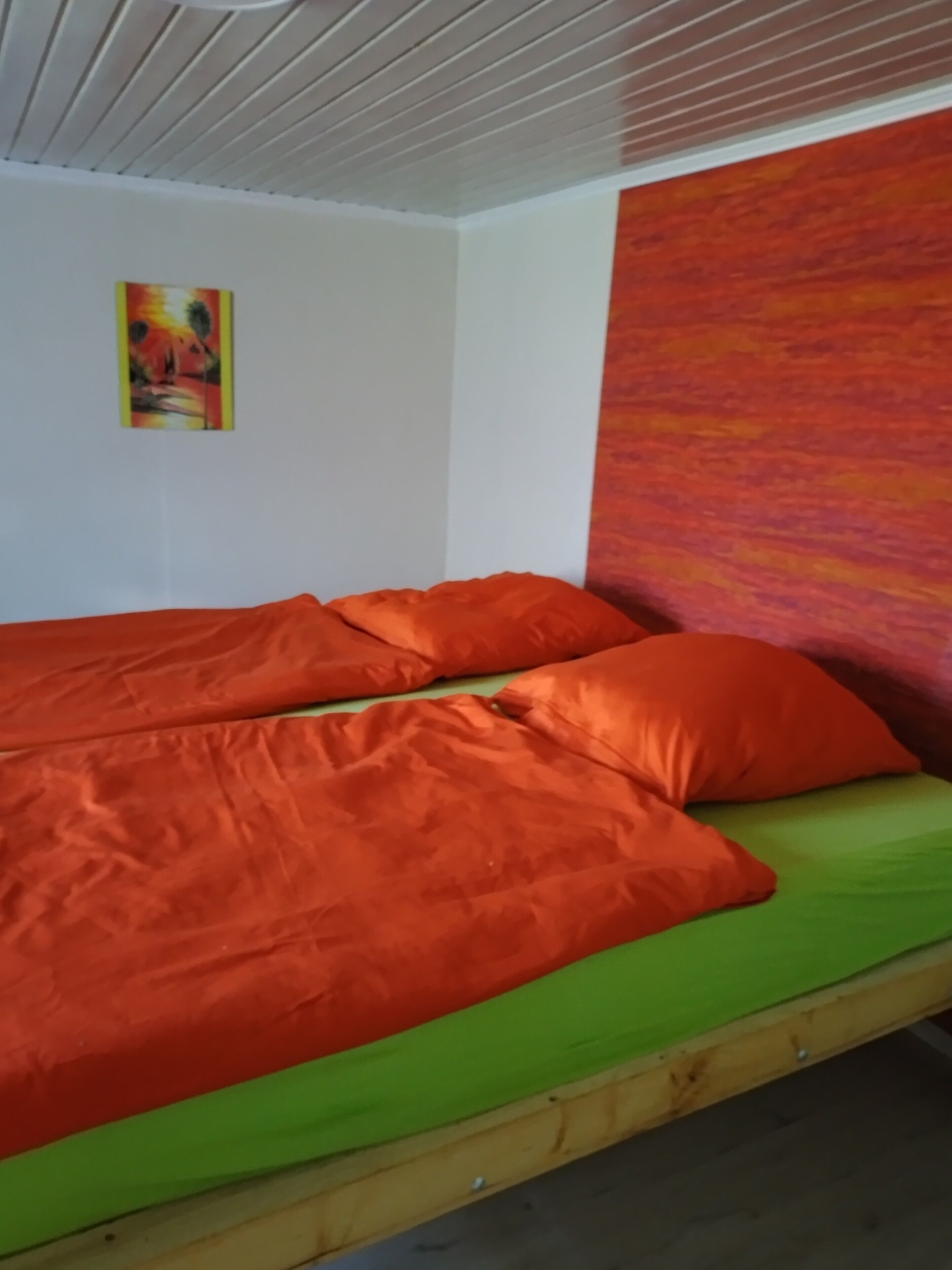 A single bed with an orange blanket and green mattress, a wooden headboard, a white ceiling, and a colorful abstract painting on the wall.