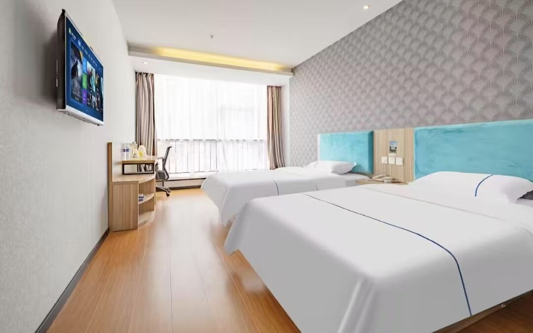 Habitación de hotel con dos camas, un televisor montado en la pared, un escritorio con una silla y una ventana con cortinas.