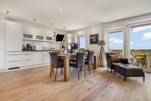 Uma cozinha moderna com armários brancos, uma área de jantar com mesa e cadeiras de madeira, e um sofá com vista para a paisagem.