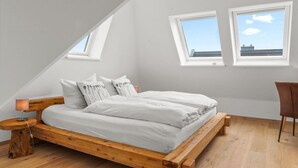 Un dormitorio con techo inclinado, estructura de cama de madera, ropa de cama blanca y dos ventanas con vistas al cielo.