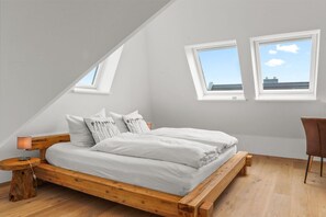 Une chambre avec un plafond en pente, un cadre de lit en bois, du linge de lit blanc et deux fenêtres donnant sur le ciel.