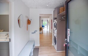 Un pasillo de hotel con un baño a la izquierda, un banco, un área de lavandería con lavadora y un comedor al fondo.