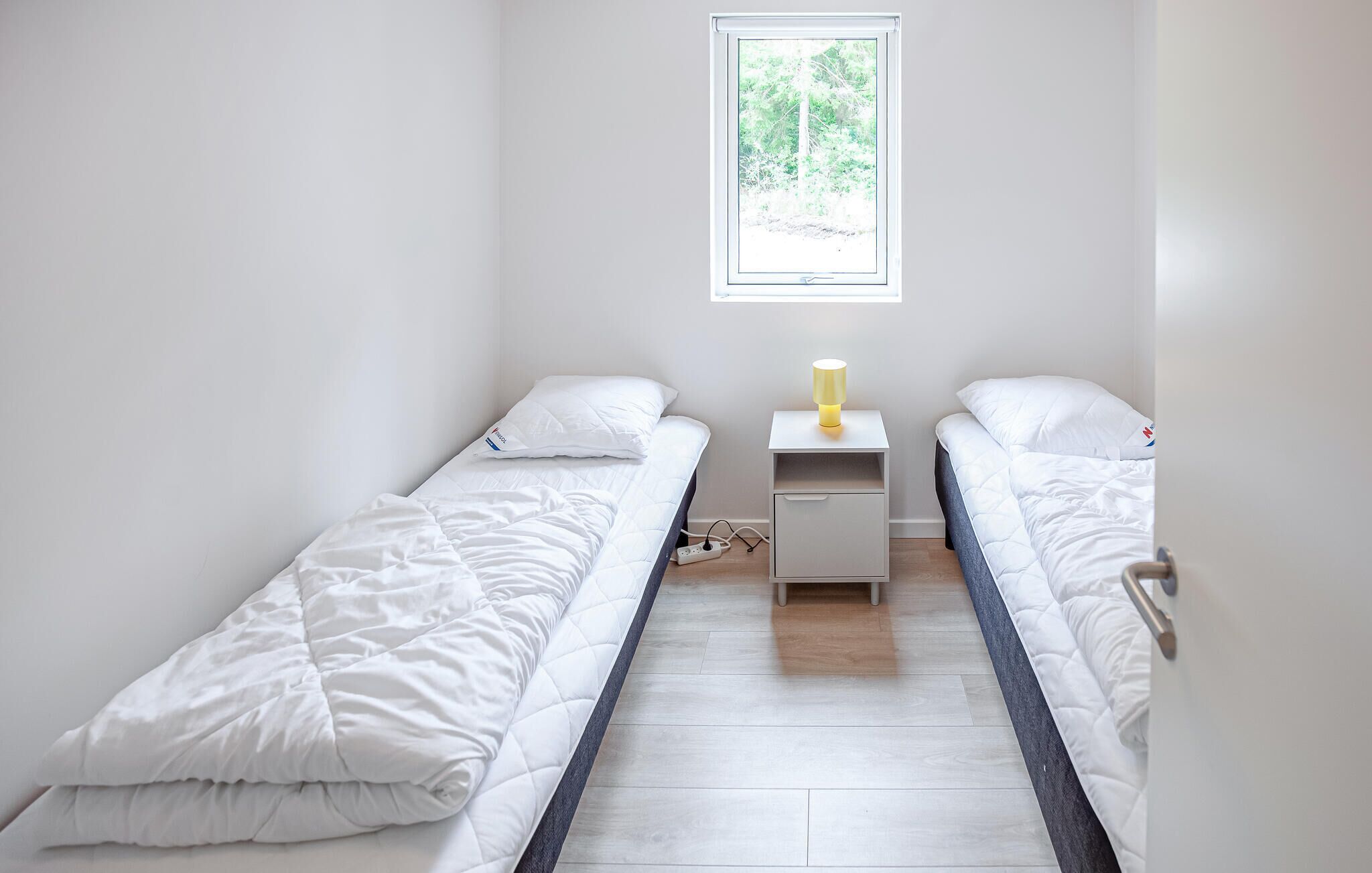 Une petite chambre propre, avec deux lits simples, une table de chevet avec une lampe et une fenêtre laissant entrer la lumière naturelle.